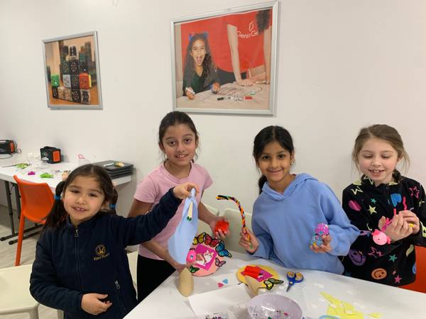 Four girls smile at the camera while showing off their colorful craft creations at a table.