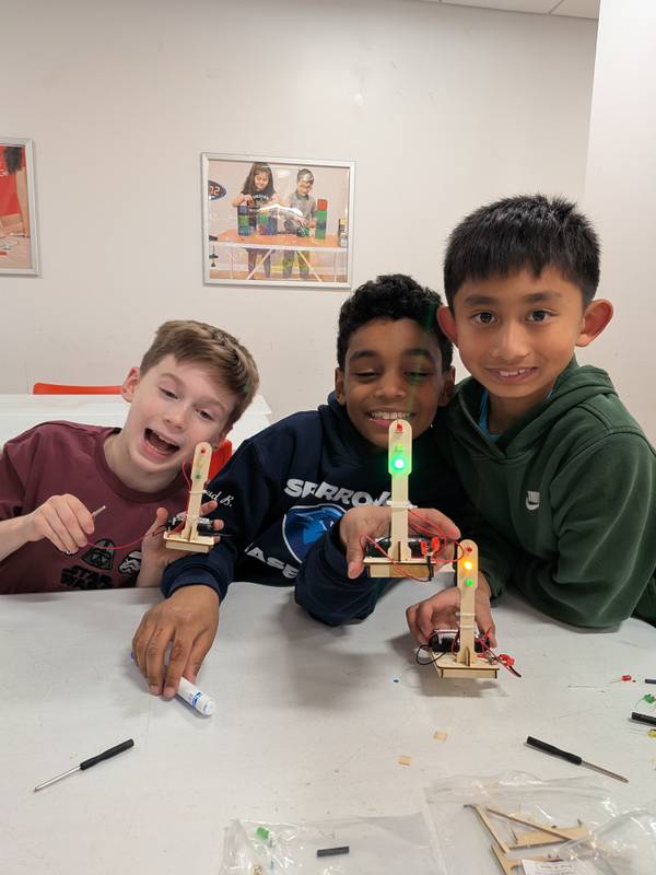 Three happy boys at a table, one with open mouth, displaying their completed traffic light circuits with glowing LEDs.