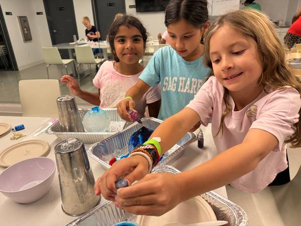 Three girls at a table excitedly engaging in a science activity involving ice, liquid dyes, and aluminum foil pans.