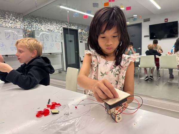 A young girl with dark hair works intently on a small wooden robot with wires on a white table, while another child works nearby.