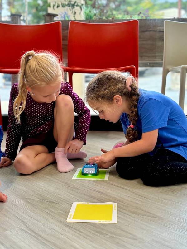 Two girls kneeling on a wood floor programming a small blue coding robot on color cards