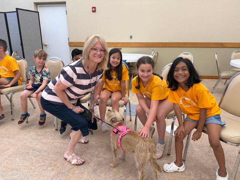 A Genius Gems staff member kneels with a small dog on a pink leash as summer campers in yellow STEM Summer Camp shirts reach out to pet it.