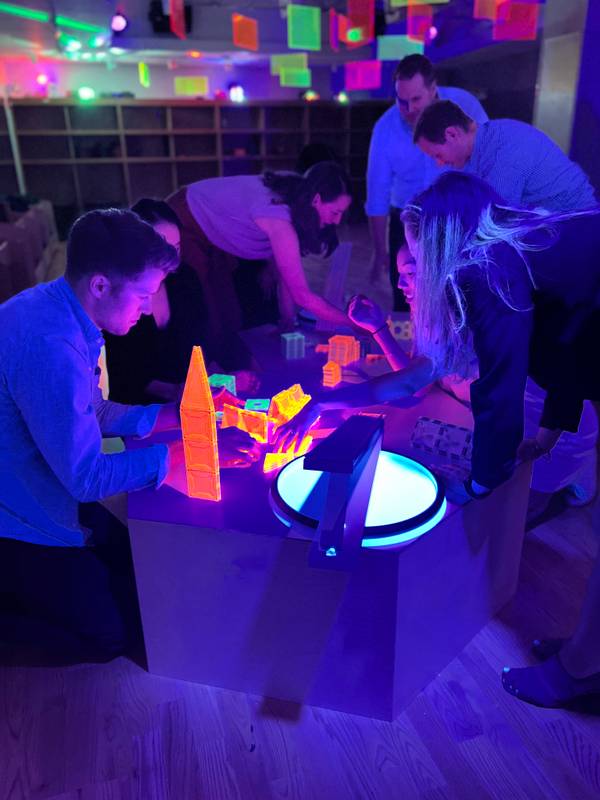Adults building glowing neon magnetic tiles around a light table under blacklight