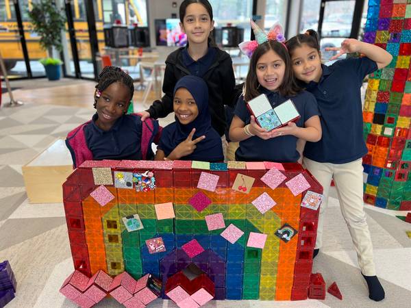 Five smiling girls in navy school uniforms behind a large rainbow magnetic-tile wall build