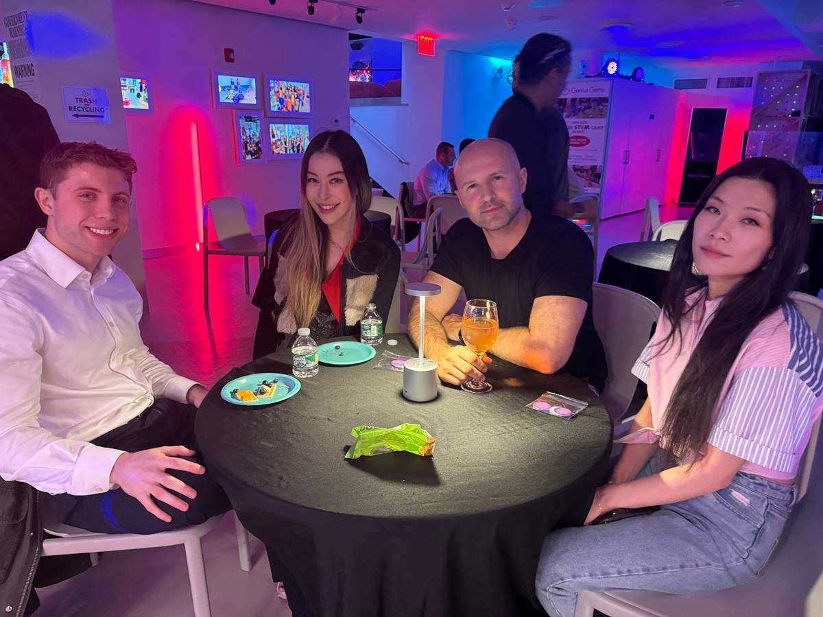 Four adults smiling around a cocktail table under pink and blue lights at an After Dark event