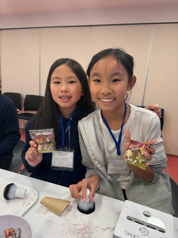Two smiling girls with school ID lanyards hold a glitter brush at a white craft table
