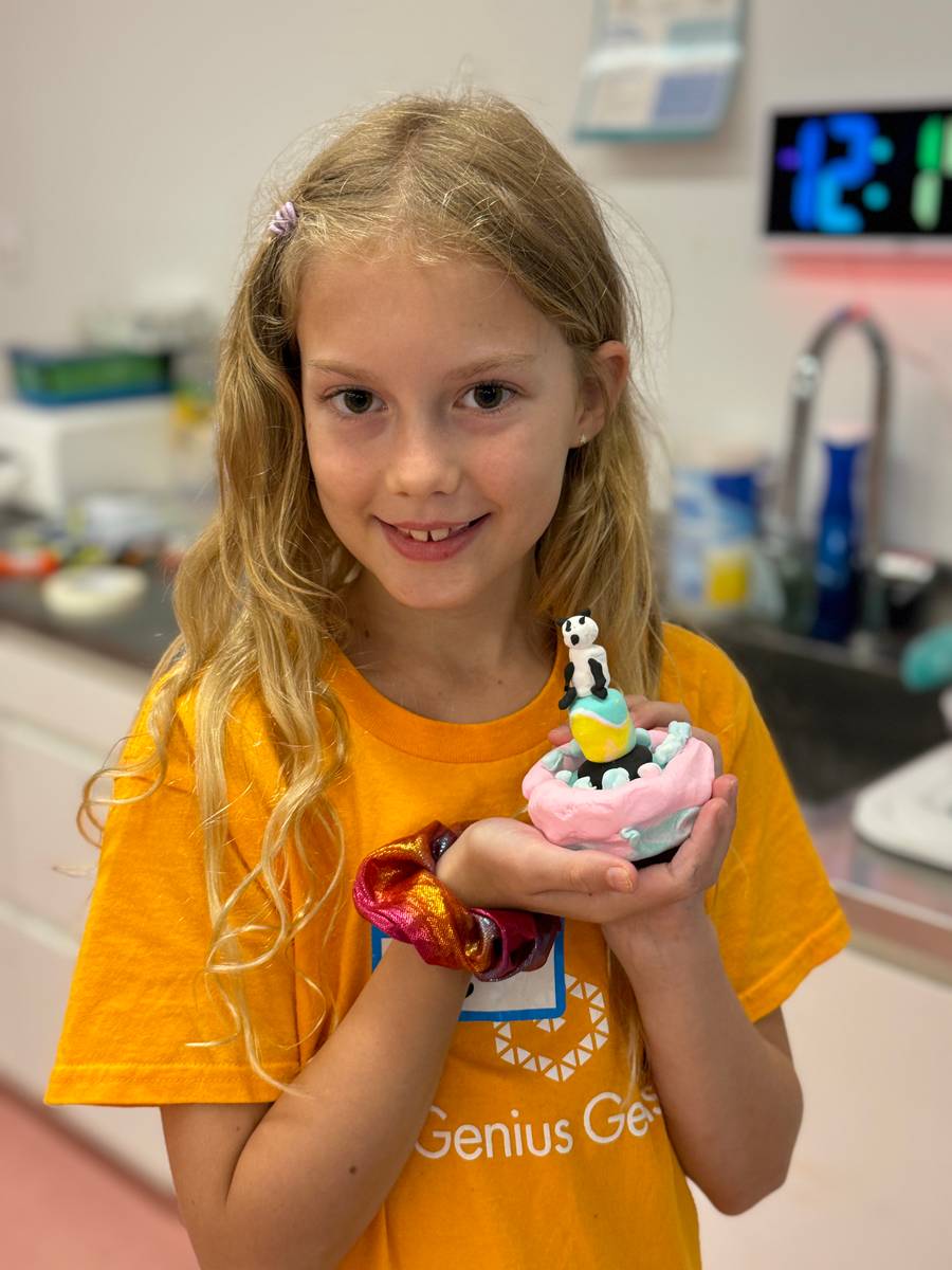 Girl in an orange Genius Gems t-shirt smiling as she holds up a clay panda she made
