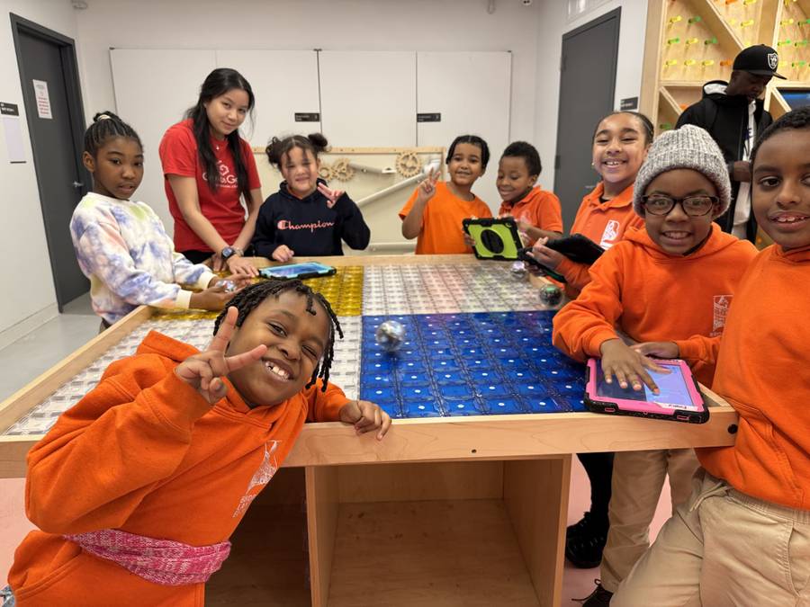 Kids in orange school shirts flash peace signs around a magnetic-tile maze table with Sphero robots and iPads