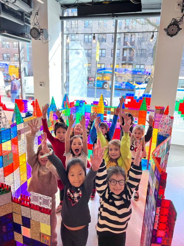 A class cheers with arms raised inside a giant rainbow magnetic-tile castle by the NYC window