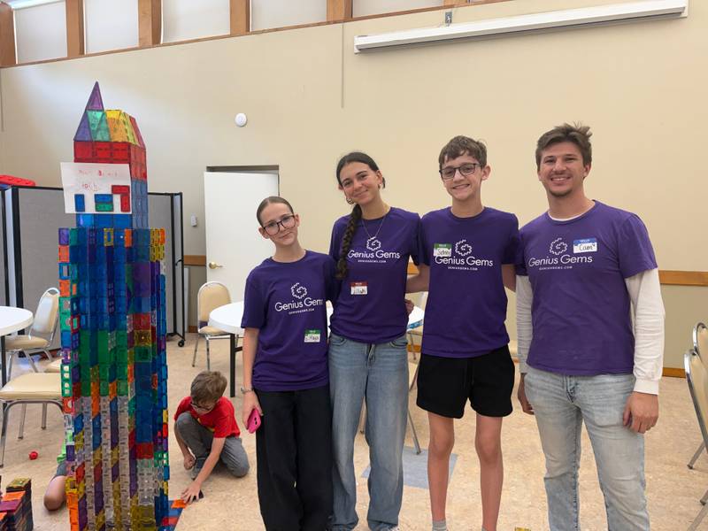 Four teen counselors-in-training in purple Genius Gems shirts stand beside a tall rainbow magnetic-tile tower in the Millburn summer camp hall while a young camper builds on the floor behind them.
