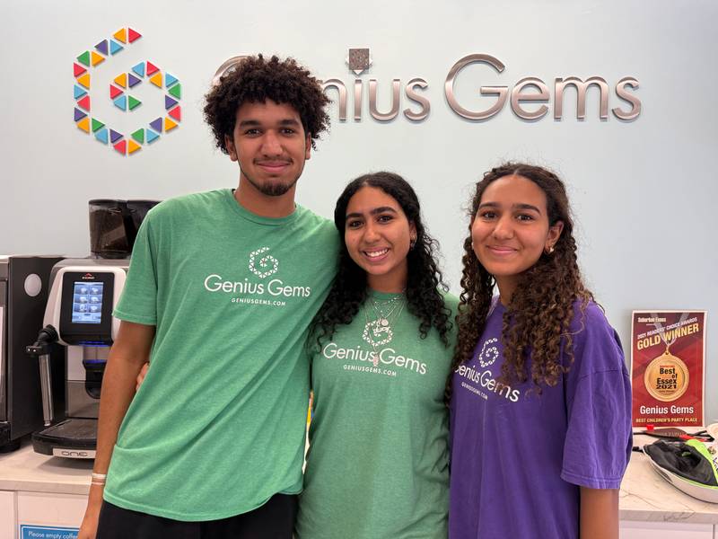 Three teen Genius Gems summer staff in green and purple team shirts smile in front of the Genius Gems logo wall at the Millburn location.