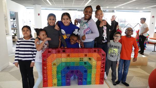 Nine kids waving from behind a rainbow tile wall