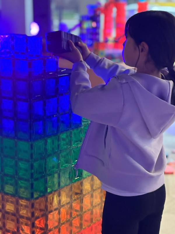 Girl in a lavender hoodie stacking magnetic tiles on top of a glowing rainbow tower