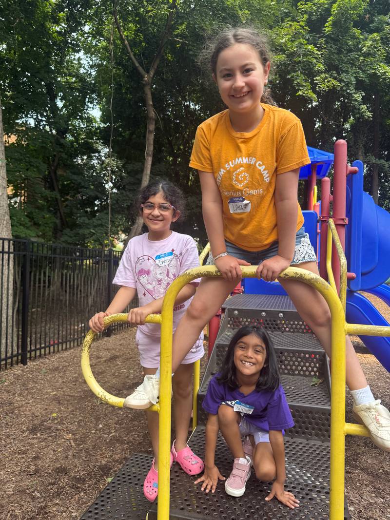 Three young Genius Gems summer campers in STEM Summer Camp shirts climb and smile on a yellow playground structure outdoors.