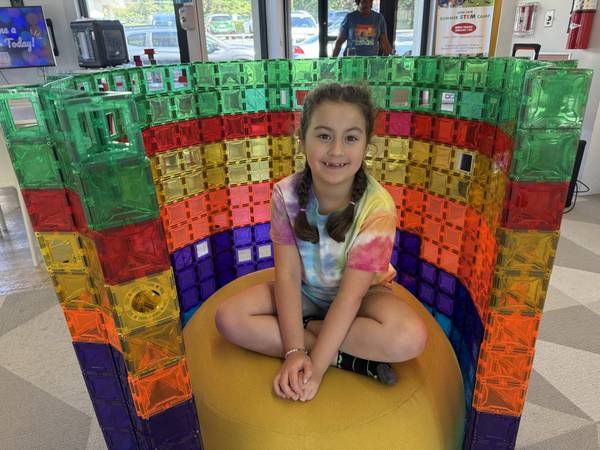 Smiling girl sitting cross-legged inside a giant rainbow magnetic-tile structure