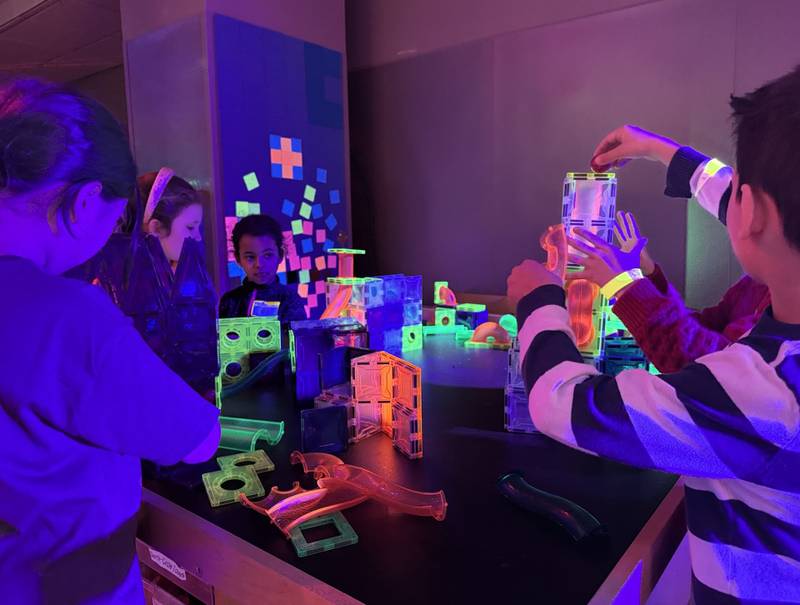 Kids building glowing magnetic-tile structures and marble runs at a blacklight table