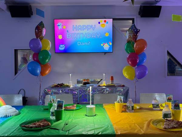 Birthday party room with colorful balloon bouquets, pizza on set tables, and a digital birthday screen.