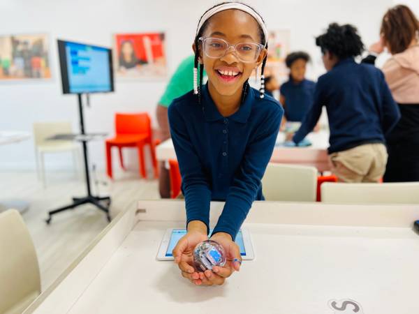 Smiling girl in a navy polo holds a clear Sphero robot ball toward the camera in a bright classroom
