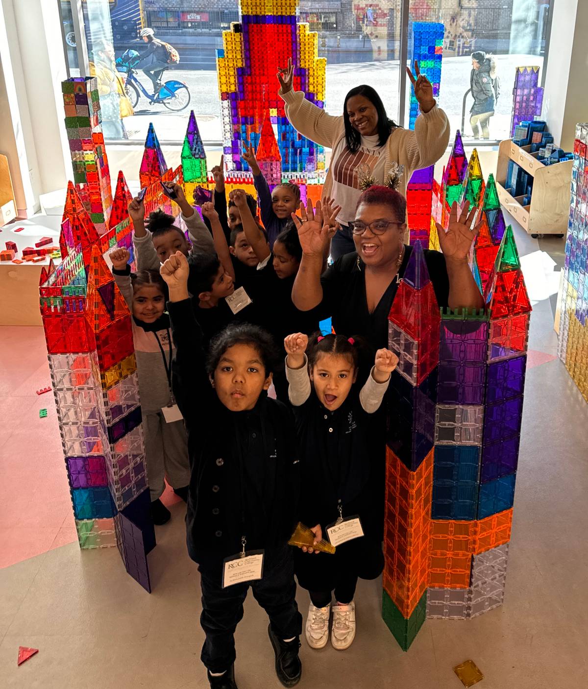 A school group and two teachers cheer with raised arms beside giant magnetic-tile towers at Genius Gems NYC