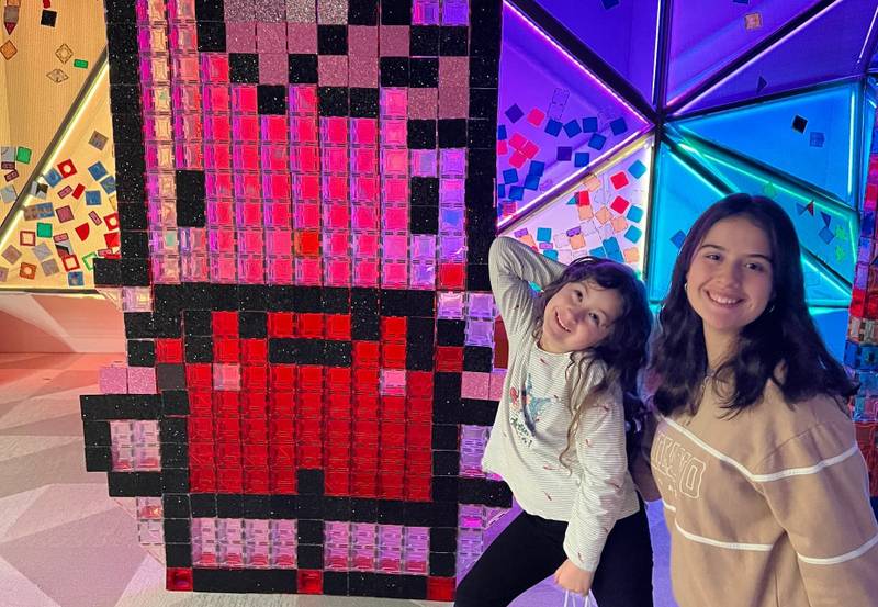 Two girls posing beside a pink and red pixel-art magnetic-tile mural
