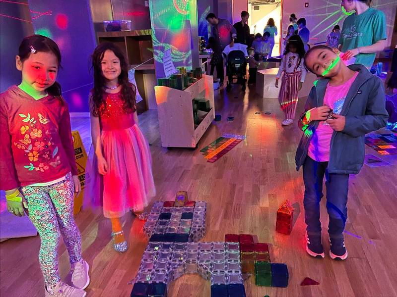 Three kids with glow sticks beside their magnetic-tile builds on the floor