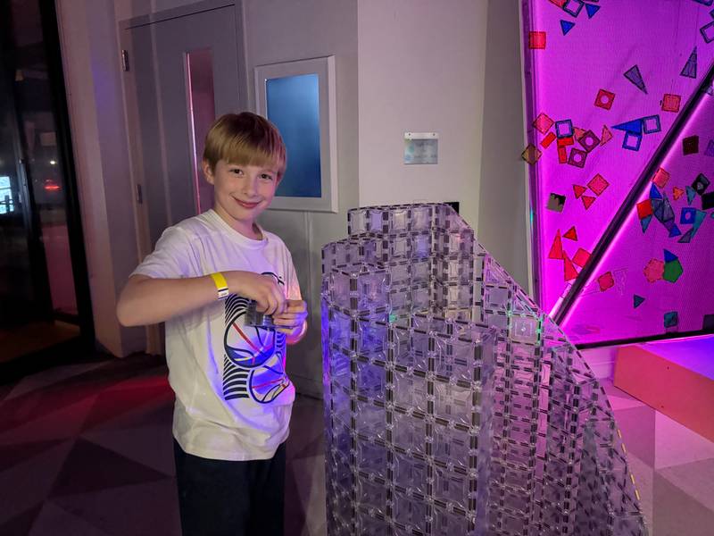 A boy adding tiles to a tall clear magnetic-tile tower under pink light