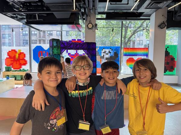 Four smiling kids on a field trip pose arm-in-arm in front of magnetic-tile pixel art murals