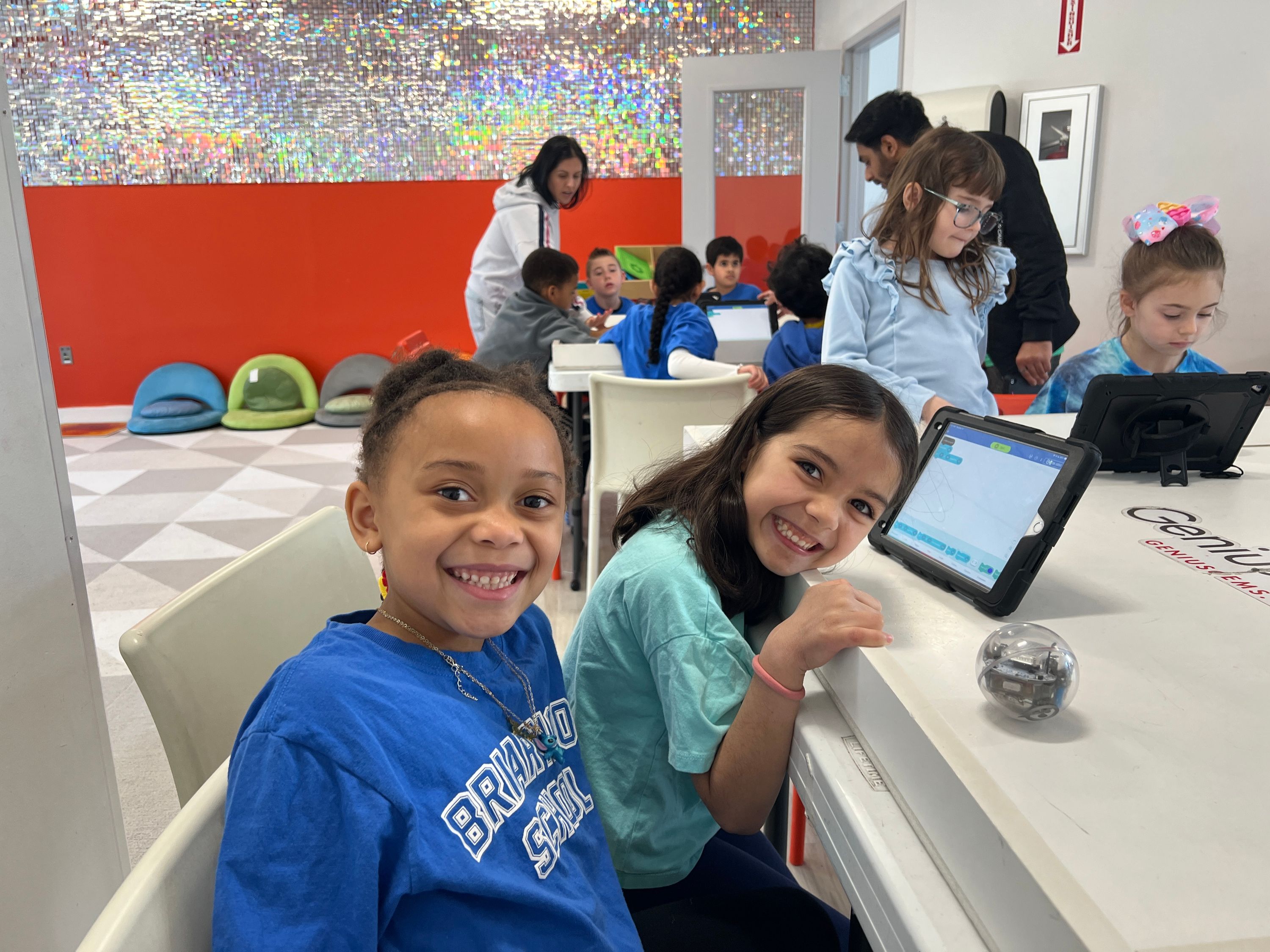 Two girls smile at the camera while coding robots at a table. Other kids and adults are visible in the bright background.