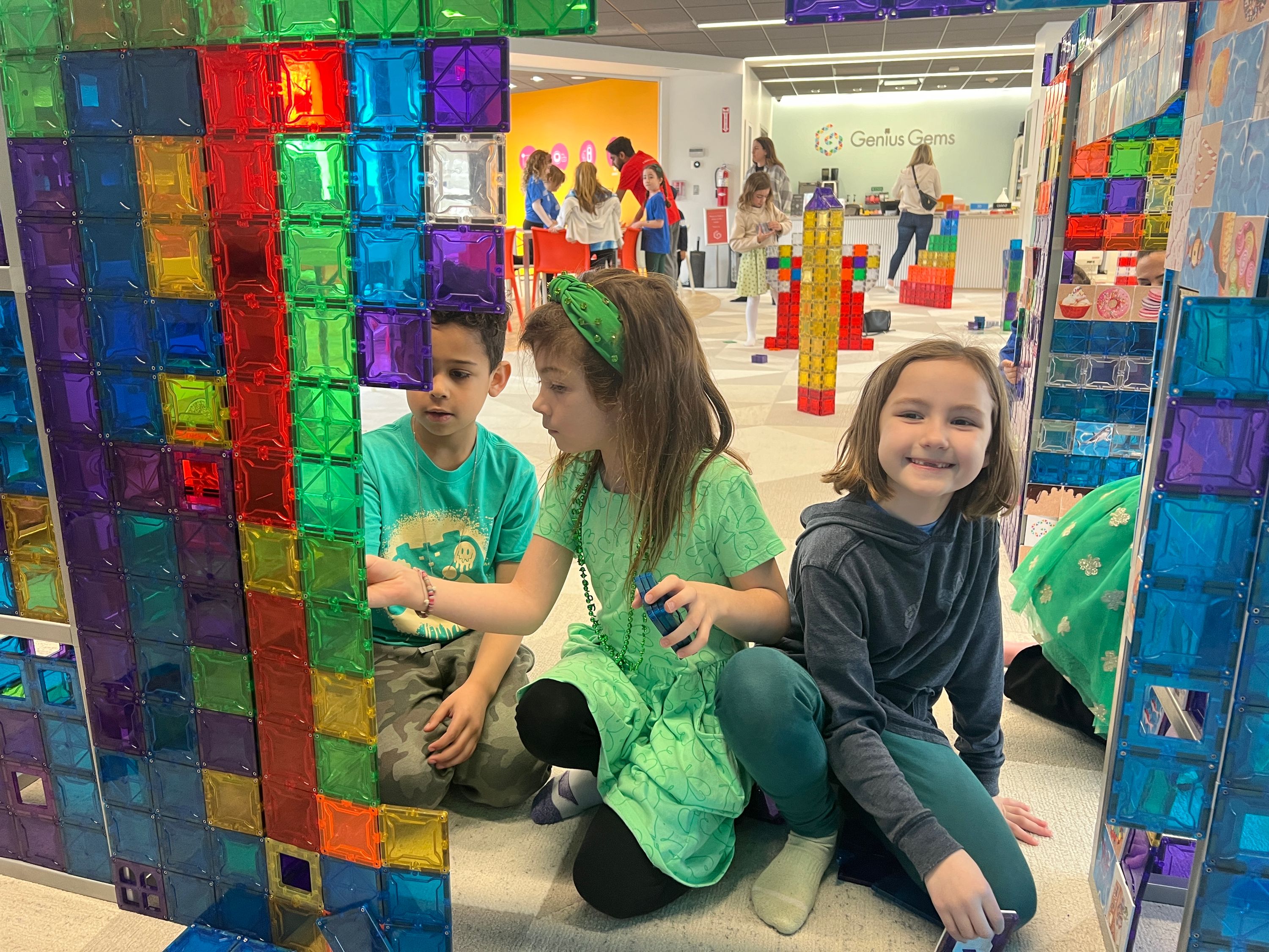 Three children are seated on the floor, constructing with colorful magnetic tiles. Behind them, other children and adults are visible.