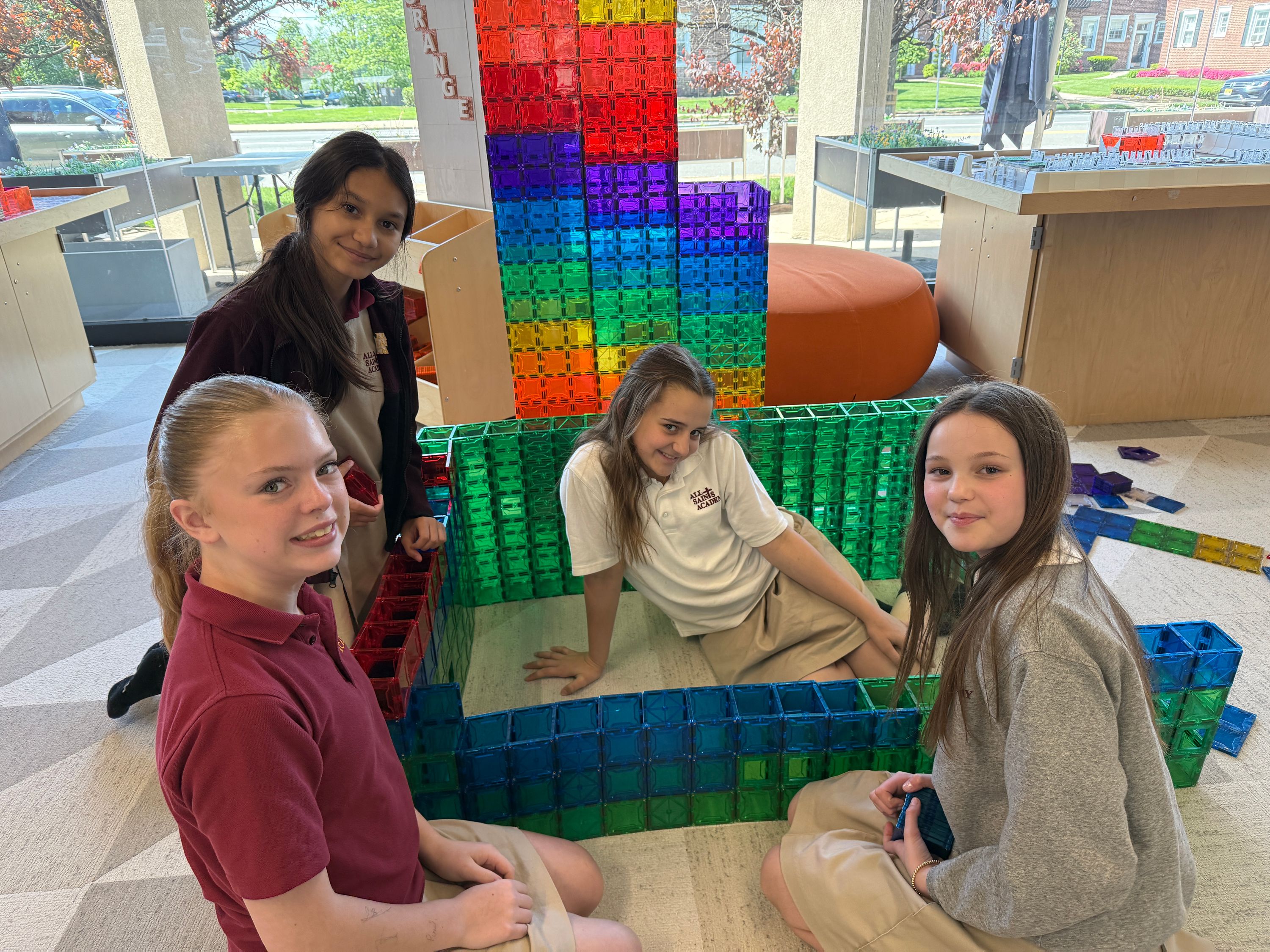 Four girls smiling inside a colorful magnetic-tile structure, a rainbow tile tower behind them